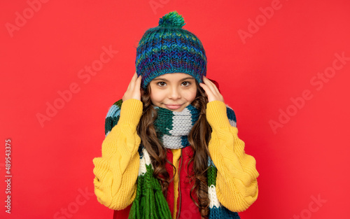 smiling teen girl in knitted winter hat and scarf feel cold on red background, cold winter