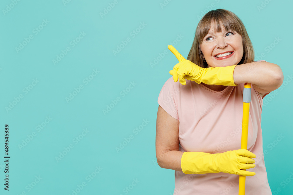 Elderly housewife woman 40s in pink t-shirt gloves doing housework leaning on mop point finger on workspace area isolated on plain pastel light blue background Housekeeping cleaning tidying up concept Elderly housewife woman 40s in pink t-shirt gloves doing housework leaning on mop point finger on workspace area isolated on plain pastel light blue background Housekeeping cleaning tidying up concept