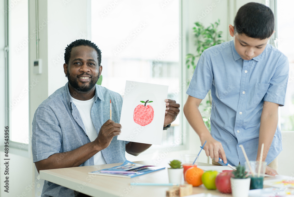 African American teacher man smiling and showing apple watercolor image of student while boy painting on paper at art school class African American teacher man smiling and showing apple watercolor image of student while boy painting on paper at art school class