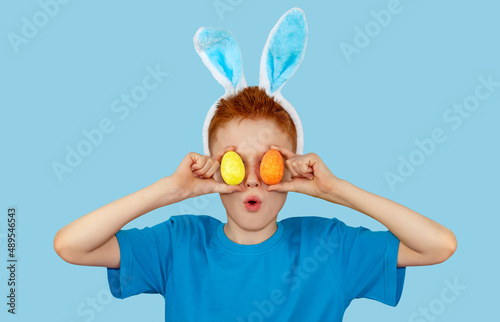 Portrait of a surprised red-haired boy in rabbit ears, who holds colorful Easter eggs in front of his eyes