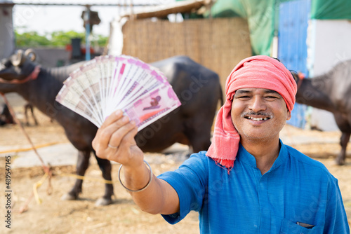 Confident milk dairy farmer showing indian currency money by looking at camera - concept of earnings, banking and business profit