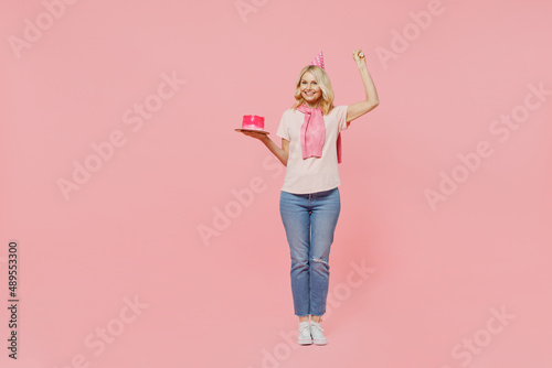 Full body elderly happy smiling cheerful fun satisfied cool woman 50s in t-shirt birthday hat hold cake do winner gesture isolated on plain pastel pink background Celebration of holiday party concept.