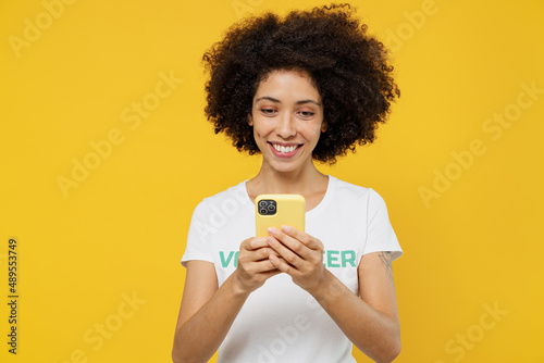 Young woman of African American ethnicity wear white volunteer t-shirt hold in hand use mobile cell phone isolated on plain yellow background Voluntary free work assistance help charity grace concept