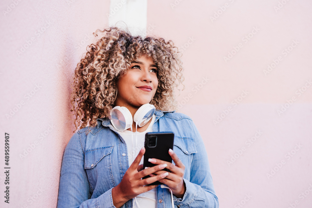© Eva - portrait of smiling hispanic woman with afro hair in city using mobile phone and headset. lifestyle
