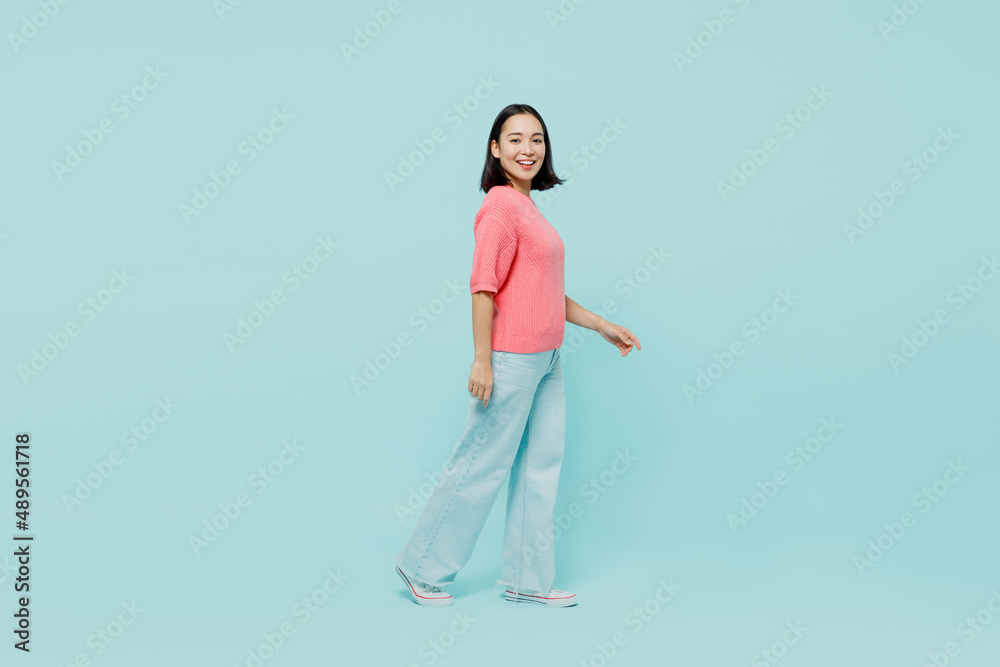 Full size young smiling happy woman of Asian ethnicity 20s wearing pink sweater walk going look camera isolated on pastel plain light blue color background studio portrait. People lifestyle concept. Full size young smiling happy woman of Asian ethnicity 20s wearing pink sweater walk going look camera isolated on pastel plain light blue color background studio portrait. People lifestyle concept.