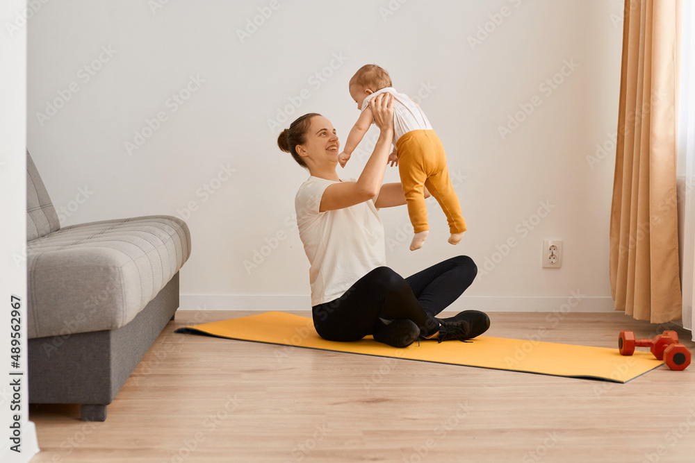 Image of attractive woman wearing white t shirt and black leggins doing sport exercises at home, sitting on mat and holding throwing up baby daughter, playing together indoor.