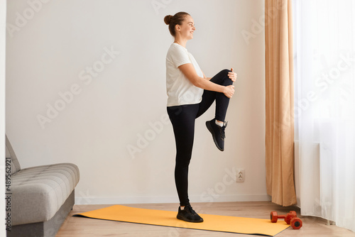 Side view full length of sporty woman wearing white t shirt and black leggins doing sport exercises at home, standing with raised leg, looking ahead, posing near sofa in front of window.