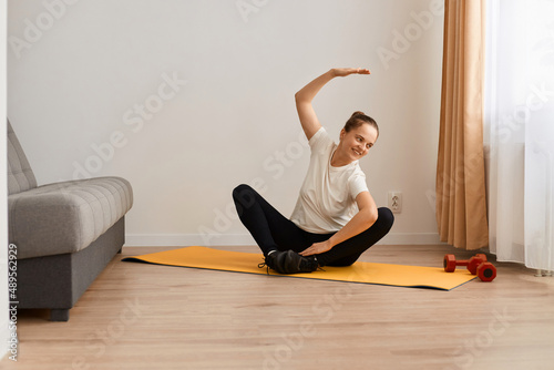 Woman wearing white t shirt and black leggins doing sport exercises at home, sitting in lotus position on mat practicing yoga, bending to side with raised hand, doing stretching workouts.