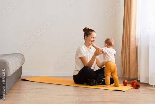 Portrait of woman young mother wearing white t shirt and black leggins doing sport exercises at home and playing with her baby daughter, sitting on mat near sofa and window.