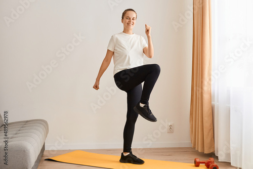 Full length portrait of woman wearing white t shirt and black leggins doing sport exercises at home, standing on mat and matching, looking smiling at camera.