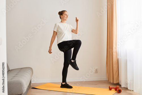 Portrait of woman wearing white t shirt and black leggins doing sport exercises at home, standing on mat and matching, looking ahead, having workout alone, healthy lifestyle.