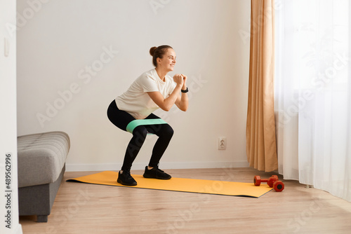 image of sporty woman wearing white t shirt and black leggins doing sport exercises at home, doing squat, lower body sport exercise, warming up and training muscles with elastic bend. © sementsova321