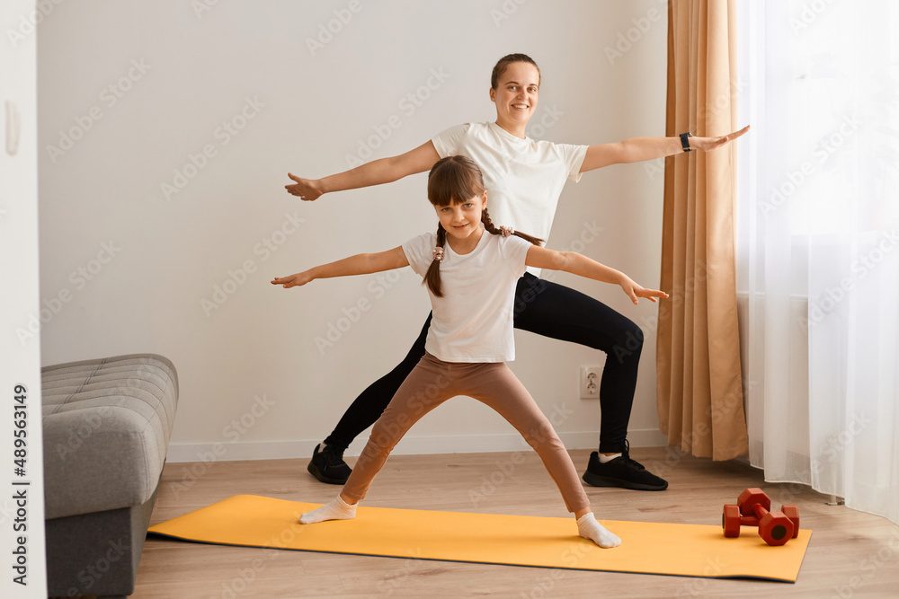 Full length portrait of woman wearing white t shirt and black leggins doing sport exercises at home with her daughter, standing in yoga pose, stretching body, training together.