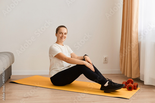 Full length shot of attractive sporty woman wearing white t shirt and black leggins doing sport exercises at home, sitting on floor and looking at camera with happy positive expression.