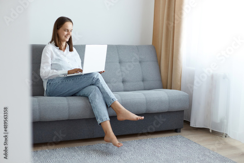 Portrait of adorable attractive woman wearing white shirt and jeans sitting on sofa and working on notebook, typing on keyboard, expressing positive emotions, enjoying online job.
