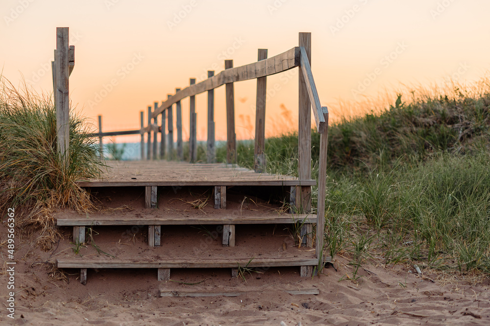 Fototapeta premium An old wooden path and stairs built over beach grass with water in the background during sunset.