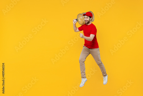 Professional side view fun delivery guy employee man in red cap T-shirt uniform workwear work as dealer courier hold water bottle jump high isolated on plain yellow background studio. Service concept.