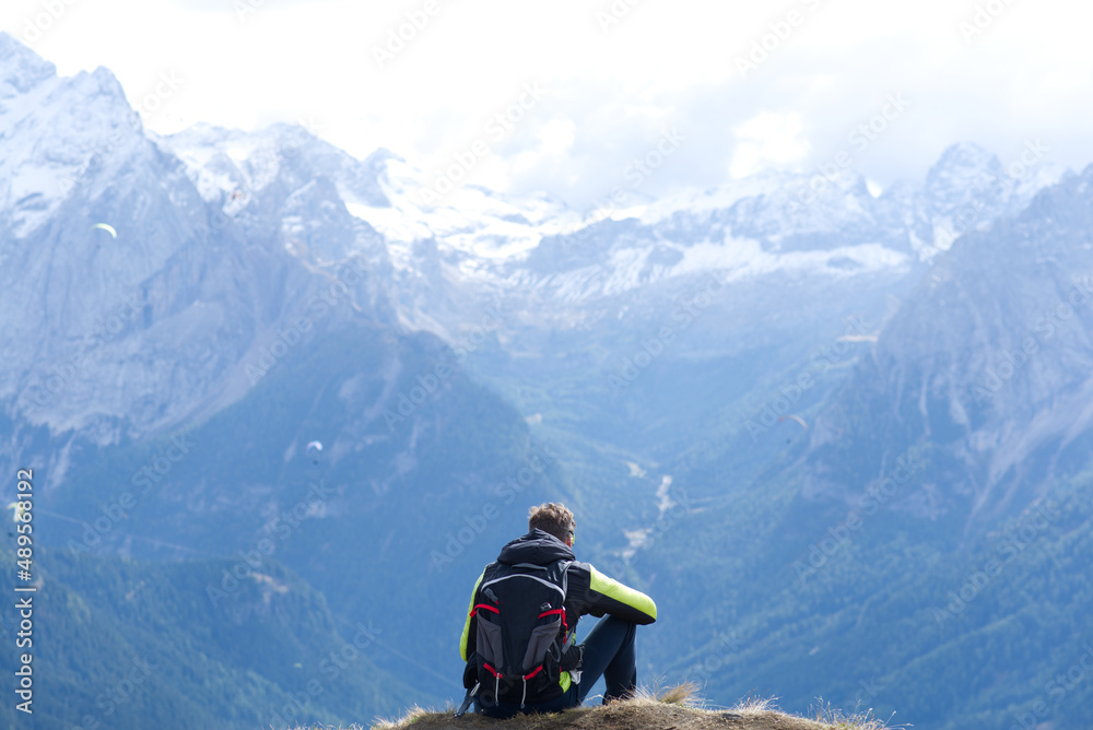 Young man with backpack sits on top of the mountain, relaxes after excursion and looks at snowy italian alps. Young man with backpack sits on top of the mountain, relaxes after excursion and looks at snowy italian alps.