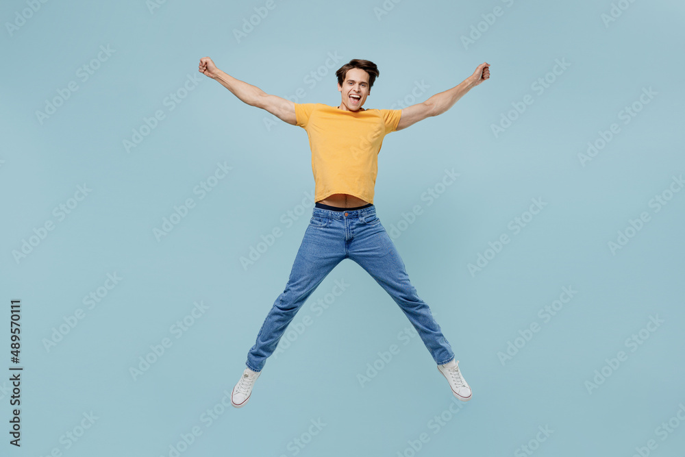 Full body overjoyed fun joyful cool young man 20s wear yellow t-shirt jump high with outstretched hands legs isolated on plain pastel light blue background studio portrait. People lifestyle concept.