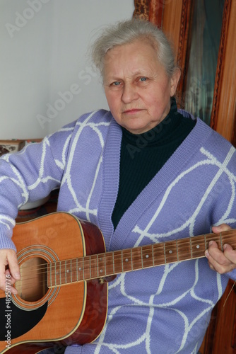 A seniour woman is playing guitar. She has blue eyes and grey hair, wears violaceous cardigan.