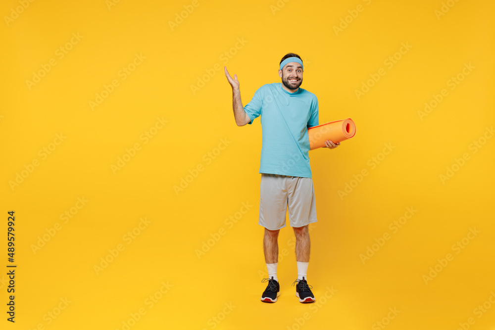 Full body young fitness trainer instructor sporty man sportsman in headband blue t-shirt spend weekend in home gym hold yoga mat spread hands isolated on plain yellow background Workout sport concept