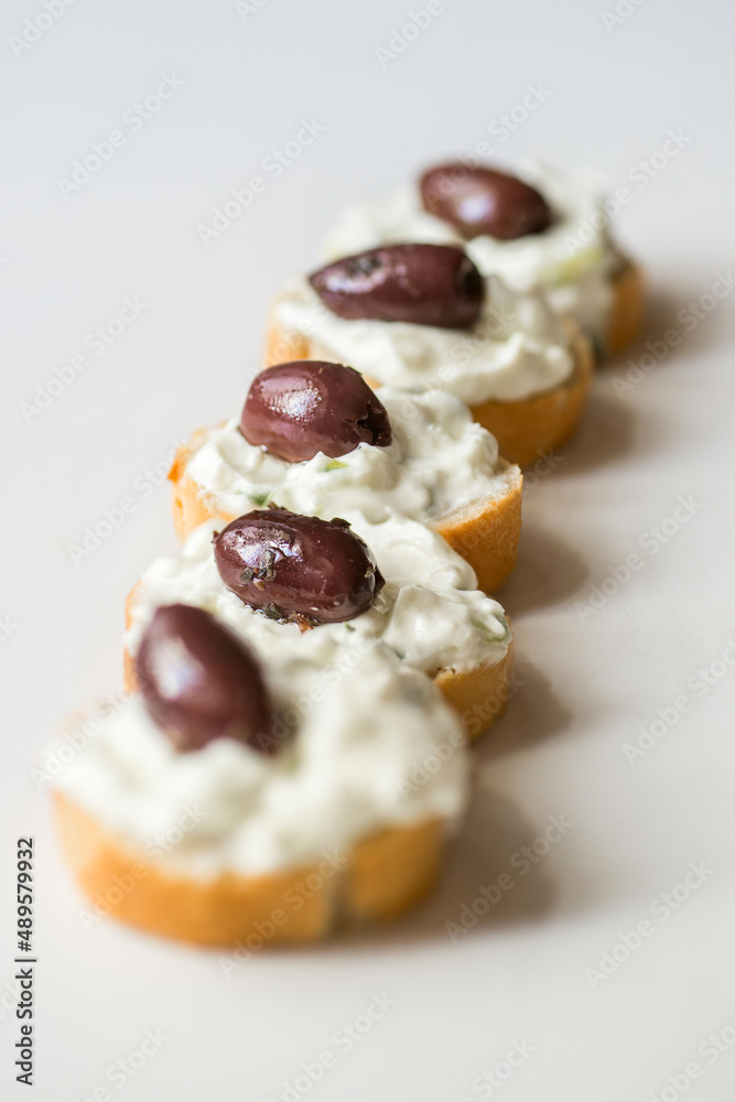 Small piece of bread with tzatziki spread and olive in row on white.