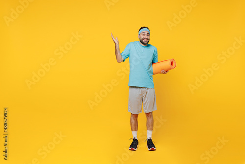 Full body young fitness trainer instructor sporty man sportsman in headband blue t-shirt spend weekend in home gym hold yoga mat spread hands isolated on plain yellow background Workout sport concept