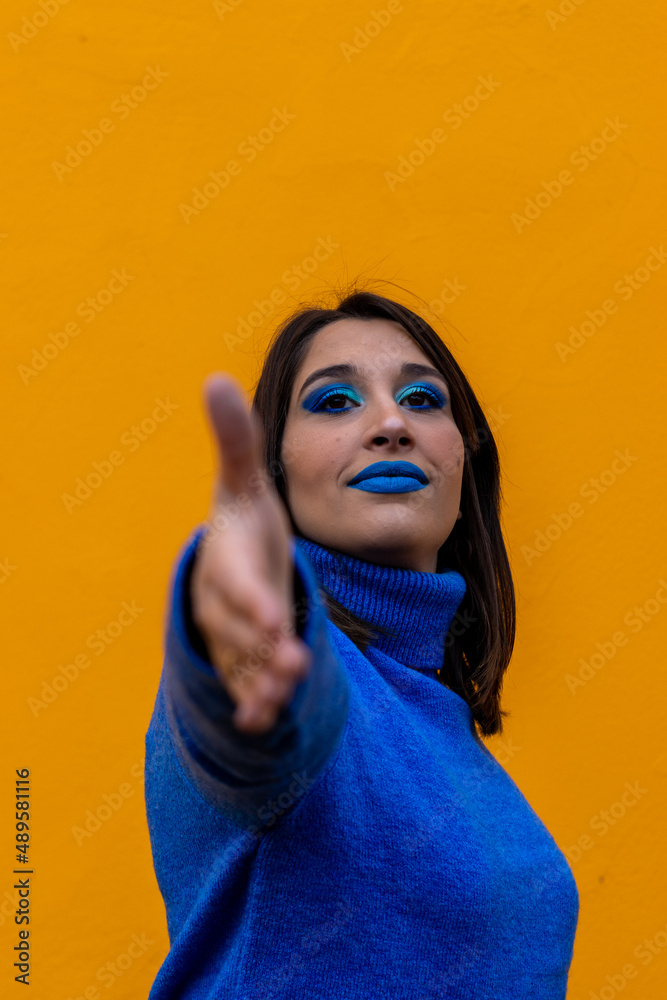 vertical portrait of caucasian woman with blue makeup and dressed in blue raising the hand to shake hands in a yellow background vertical portrait of caucasian woman with blue makeup and dressed in blue raising the hand to shake hands in a yellow background