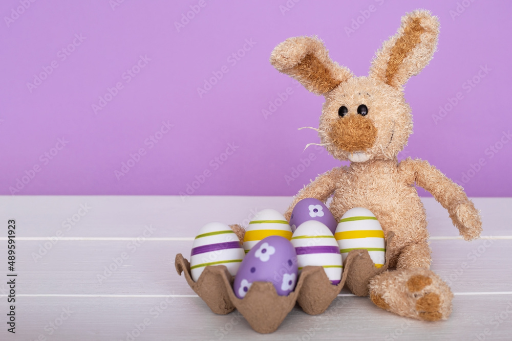 Fototapeta premium Easter composition - a rabbit and colorful eggs on a lilac background.