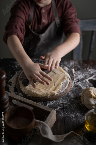 Kid making pelmeni (meet dumplings) of dough on wooden table with ingredients flour, oil, salt, dark background. Copy space. Home bakery concept, kitchen cooking story. © Diana