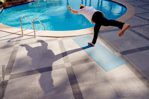 Asana standing on one leg, meditation yoga caucasian man in white shirt on background pool, palm trees and sunlight © Parilov