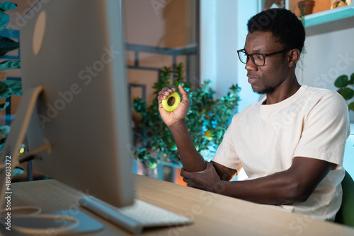 Tablou pe pânză Young African man freelancer exercising with silicone grip ring at cozy home office, remote worker doing hand exercises while resting from computer at workplace