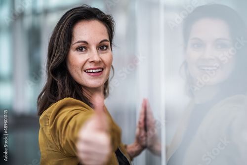 happy smiling casual brunette businesswoman with occa mustard jacket and thumb up in a optimistic and motivating pose sourrounded by a modern office background