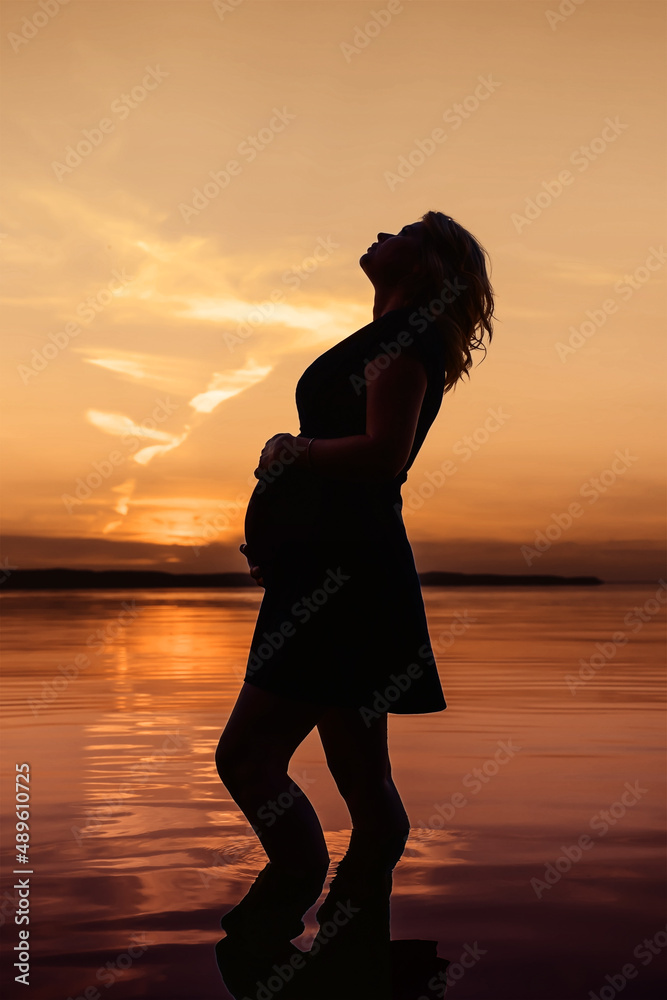 Full length silhouette unrecognized female expecting a baby, who stands in the water of the lake and touch her belly against evening sky background at sunset. Pregnancy, vertical, toned image 