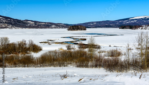 Winter landscape with a tributary River entering the frozen lake with with lake island and hills in the background background. Frozen Lake on a cold winter day. 