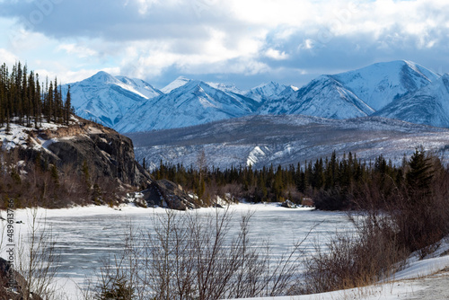 Photography bright blue frozen winter scene of ice covered lake with snow, tree line, mountains and very cloudy overcast sky