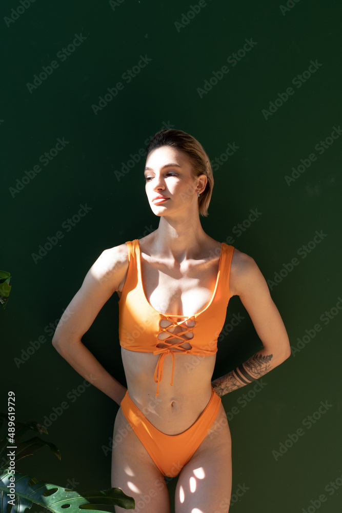 Obraz premium young woman posing in an orange swimsuit in the studio, standing near a green wall