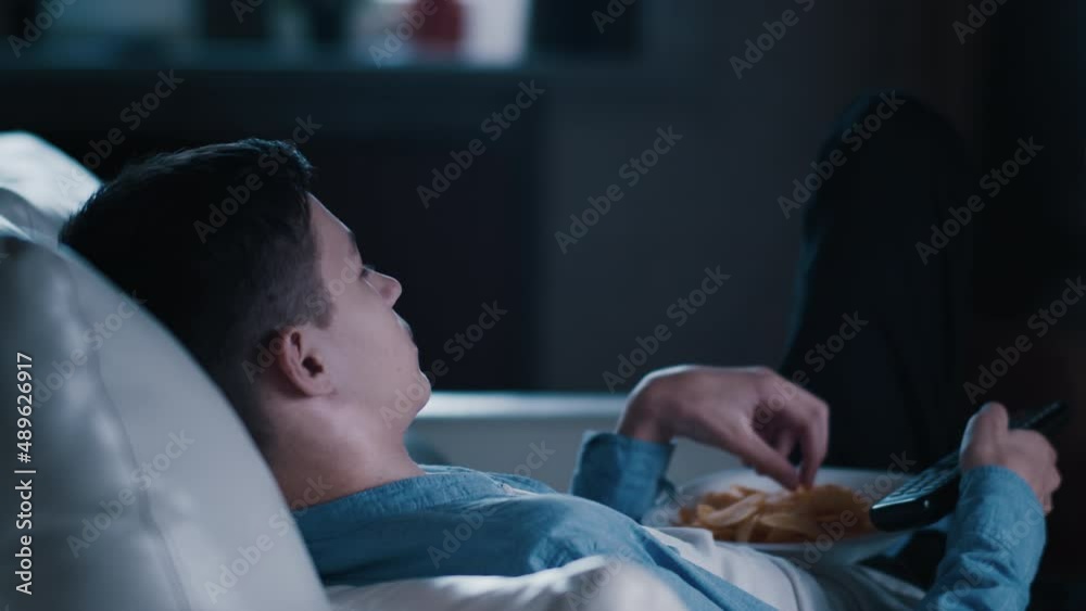 teen boy lies on the sofa watching TV at night at home and eating potato chips, switching channels, relax on sofa teen boy lies on the sofa watching TV at night at home and eating potato chips, switching channels, relax on sofa