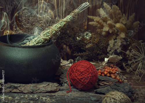 Magic items. Smoking twist of bitter wormwood herb for fumigation in iron cauldron, a ball of red wool, pine bark and dry flowers on wooden background, dark key, selective focus. photo