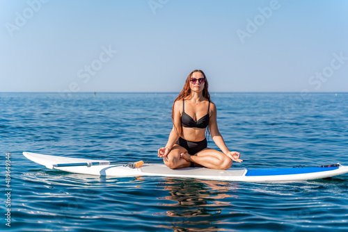 Sporty girl on a glanders surfboard in the sea on a sunny summer day. In a striped swimsuit, he sits in the lotus position with his eyes closed. © svetograph
