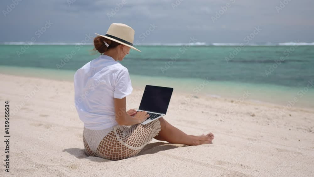Successful business woman in yellow hat working on laptop by the sea. Pretty lady typing on computer at summer day outdoors. Freelance, travel and holidays concept. Successful business woman in yellow hat working on laptop by the sea. Pretty lady typing on computer at summer day outdoors. Freelance, travel and holidays concept.