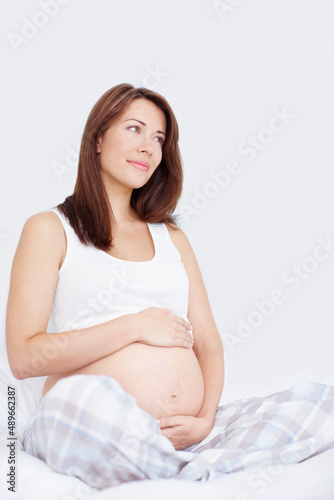 Expectations and dreams. Pretty young woman sitting on her bed holding her bare pregnant belly while looking away in thought. © Stigur/peopleimages.com