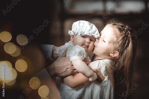 Vászonkép the elder sister kisses a baby in a baptismal outfit in a temple or church who c