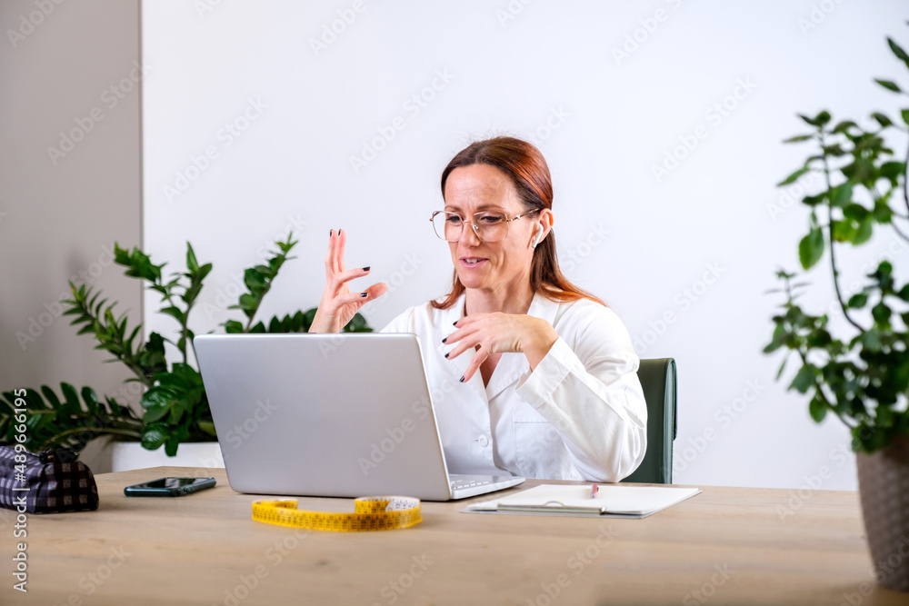 Portrait of female nutritionist in medical gown using laptop for online consultation via video chat with her patient while sitting in her office. Dietitian with laptop giving online weight loss Portrait of female nutritionist in medical gown using laptop for online consultation via video chat with her patient while sitting in her office. Dietitian with laptop giving online weight loss