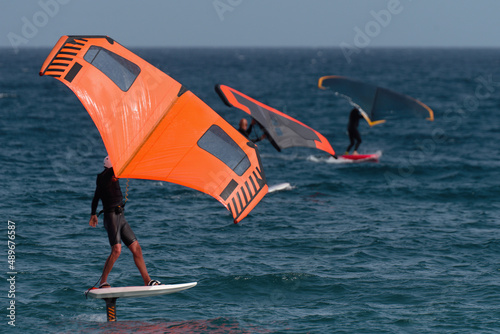 A group is wing foiling using handheld inflatable wings and hydrofoil surfboards in a blue ocean, rider on a wind wing board, surf the waves © pavel1964