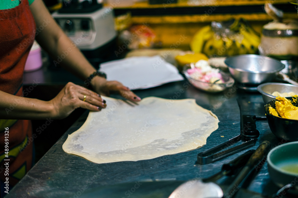 Fototapeta premium Cooking sweet Thai pancakes. A woman on the street rolls and stretches the dough straightens a close-up of her hand. traditional Asian Thai street food, lifestyle. food for tourists