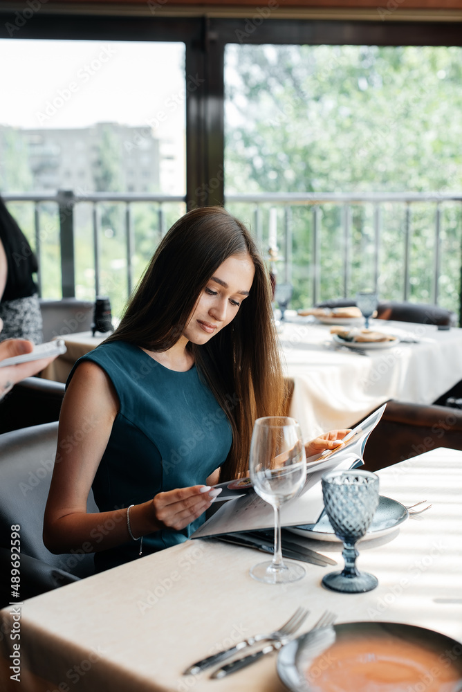 A young beautiful woman in an elegant restaurant looks through the menu and makes an order to a young waiter in a stylish apron. Customer service in the restaurant and catering establishments. A young beautiful woman in an elegant restaurant looks through the menu and makes an order to a young waiter in a stylish apron. Customer service in the restaurant and catering establishments.