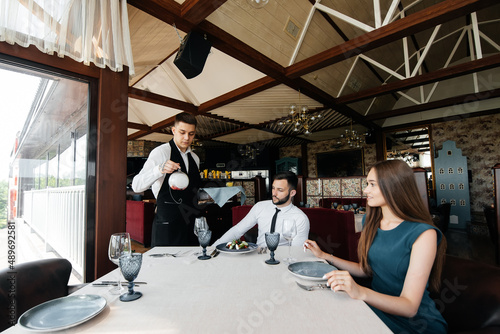 A young waiter in a stylish apron serves a table with a beautiful couple in an elegant restaurant. Customer service in an elite restaurant and public catering establishment.