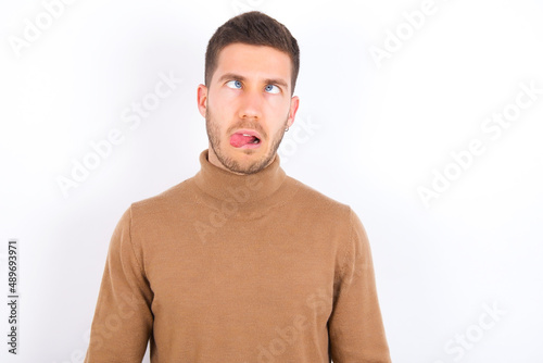 young caucasian man wearing grey turtleneck over white background showing grimace face crossing eyes and showing tongue. Being funny and crazy © Roquillo