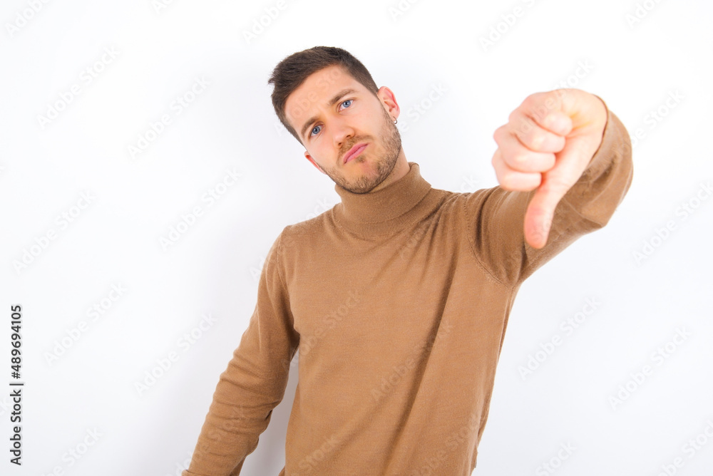 young caucasian man wearing grey turtleneck over white background feeling angry, annoyed, disappointed or displeased, showing thumbs down with a serious look